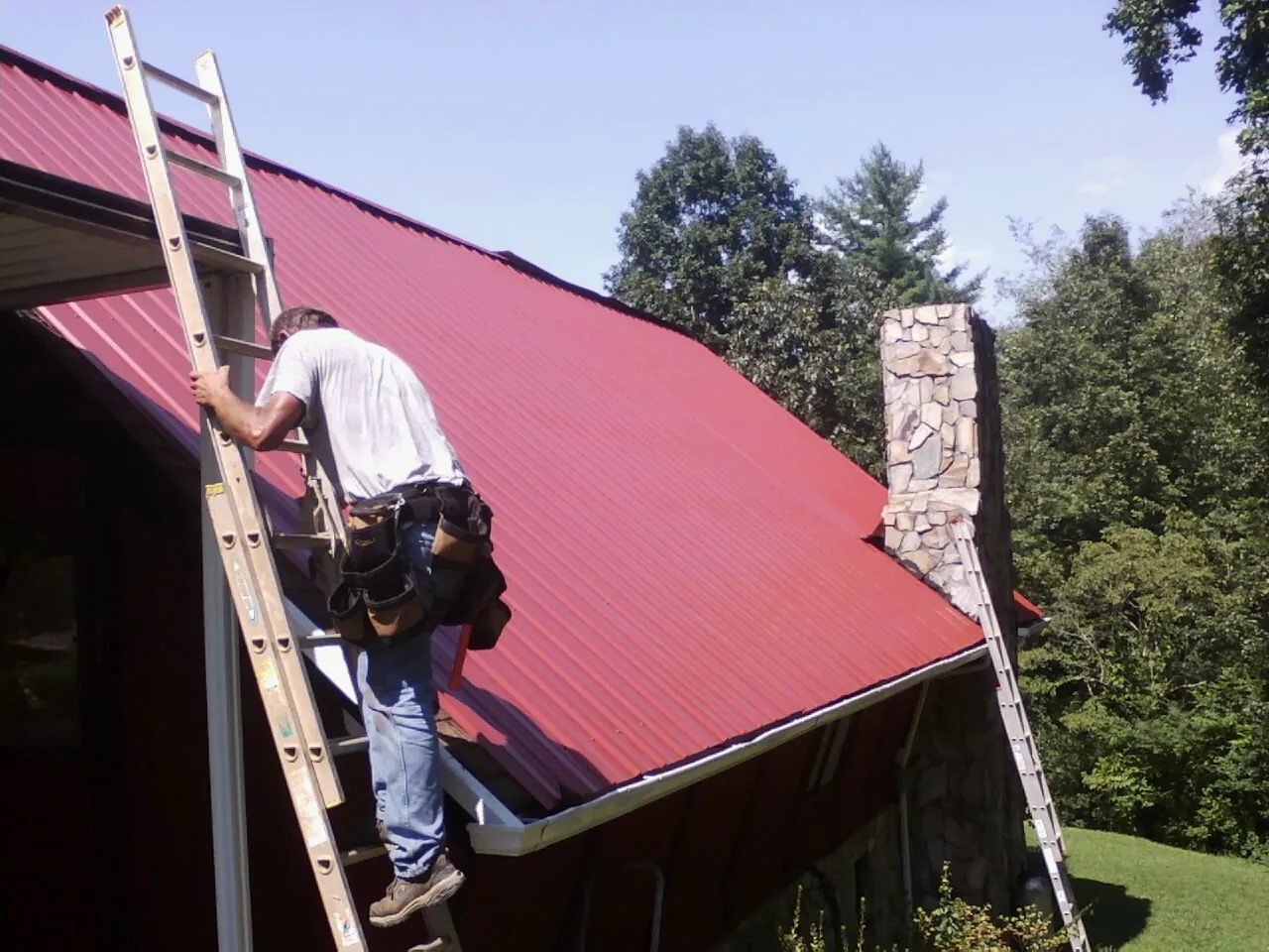 Roofer on a ladder beside a red metal roof and stone chimney — Soffit & Fascia Repair by Roofing Brothers in Wales